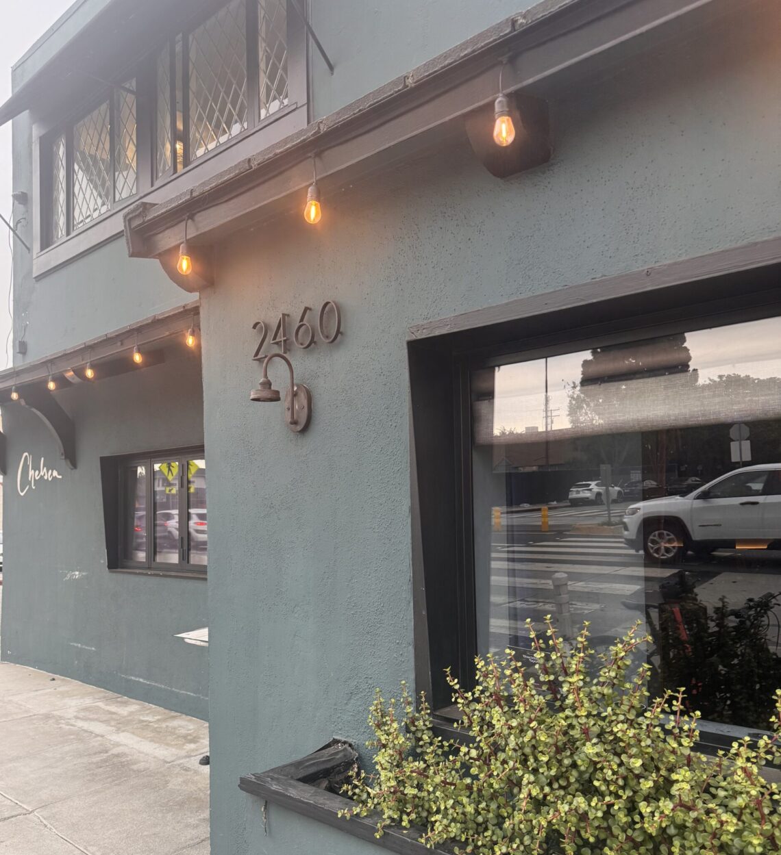 A street-level, angled view of the exterior of Chelsea Restaurant. The two-story building features a matte, dark slate-gray stucco facade. On the main corner wall, the house number "2460" is mounted in dark metal above a matching gooseneck light fixture. Warm, amber bistro string lights hang under the roofline eaves. A large glass window reflects the street outside, while a black planter box beneath it overflows with small-leafed green succulents. Further down the sidewalk, the word "Chelsea" is written in elegant, white cursive script on the building's wall.