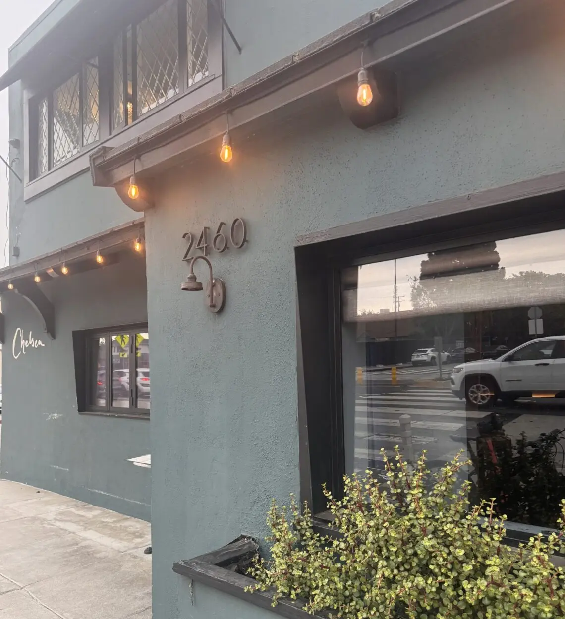 A street-level, angled view of the exterior of Chelsea Restaurant. The two-story building features a matte, dark slate-gray stucco facade. On the main corner wall, the house number "2460" is mounted in dark metal above a matching gooseneck light fixture. Warm, amber bistro string lights hang under the roofline eaves. A large glass window reflects the street outside, while a black planter box beneath it overflows with small-leafed green succulents. Further down the sidewalk, the word "Chelsea" is written in elegant, white cursive script on the building's wall.