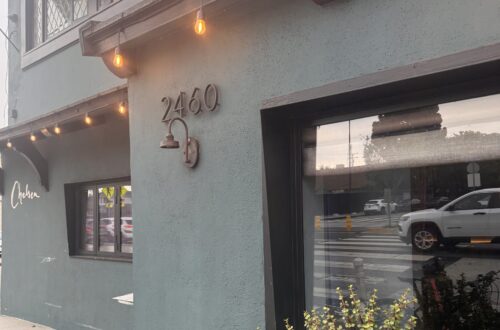 A street-level, angled view of the exterior of Chelsea Restaurant. The two-story building features a matte, dark slate-gray stucco facade. On the main corner wall, the house number "2460" is mounted in dark metal above a matching gooseneck light fixture. Warm, amber bistro string lights hang under the roofline eaves. A large glass window reflects the street outside, while a black planter box beneath it overflows with small-leafed green succulents. Further down the sidewalk, the word "Chelsea" is written in elegant, white cursive script on the building's wall.