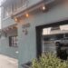 A street-level, angled view of the exterior of Chelsea Restaurant. The two-story building features a matte, dark slate-gray stucco facade. On the main corner wall, the house number "2460" is mounted in dark metal above a matching gooseneck light fixture. Warm, amber bistro string lights hang under the roofline eaves. A large glass window reflects the street outside, while a black planter box beneath it overflows with small-leafed green succulents. Further down the sidewalk, the word "Chelsea" is written in elegant, white cursive script on the building's wall.