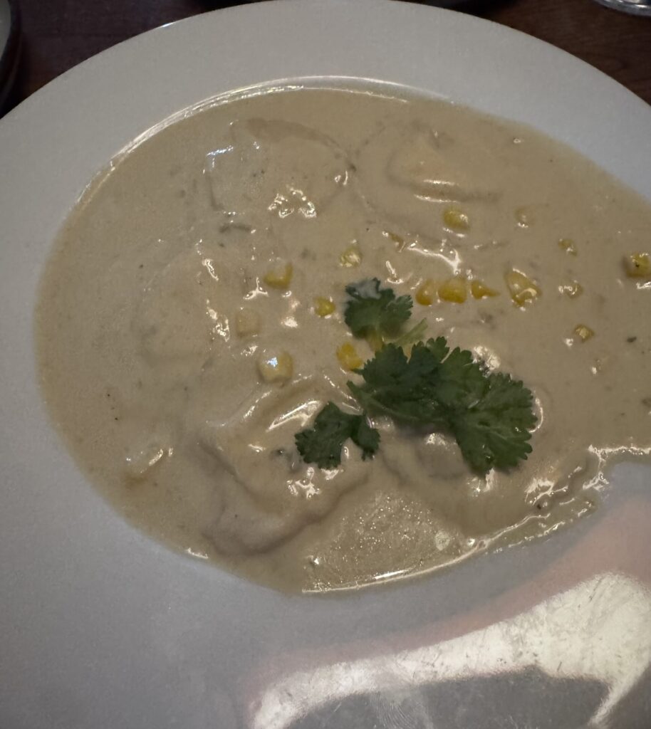 A top-down shot of a pasta dish served in a wide, shallow white bowl on a dark wood restaurant table. Several pillows of ravioli pasta are completely submerged under a thick, creamy, light-beige Thai green curry sauce. The dish is garnished with scattered bright yellow sweet corn kernels and a single fresh green cilantro sprig resting right in the center.