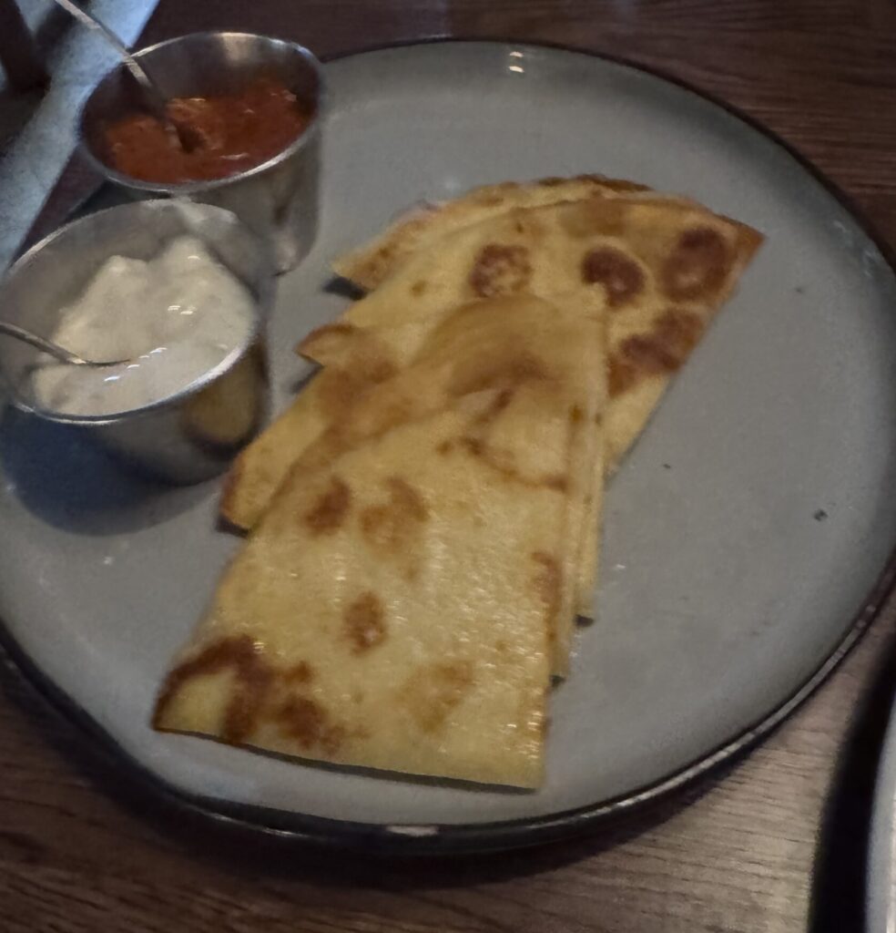 A top-down shot of a light blue round plate on a dark wooden table in a restaurant setting. On the plate, several folded pieces of warm, golden-brown naan bread are neatly stacked. Next to the bread are two small metal ramekins: one filled with a creamy white raita sauce, and the other filled with a vibrant red chili sauce, each with a small spoon resting inside.