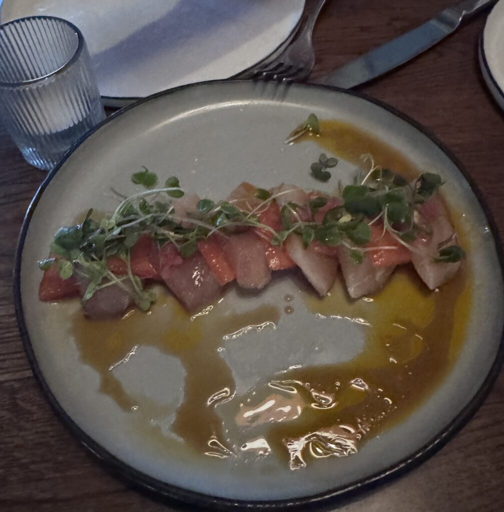 A top-down shot of a beautifully plated appetizer on a round gray ceramic plate with a dark rim, resting on a wooden restaurant table. Alternating slices of pale pink yellowtail fish and vibrant red watermelon sashimi are neatly arranged in a gentle curve across the center. The dish is pooled in a light brown ponzu and golden-orange chili oil sauce, topped with thinly sliced green serrano peppers, finely diced onions, and a delicate row of fresh microgreens.