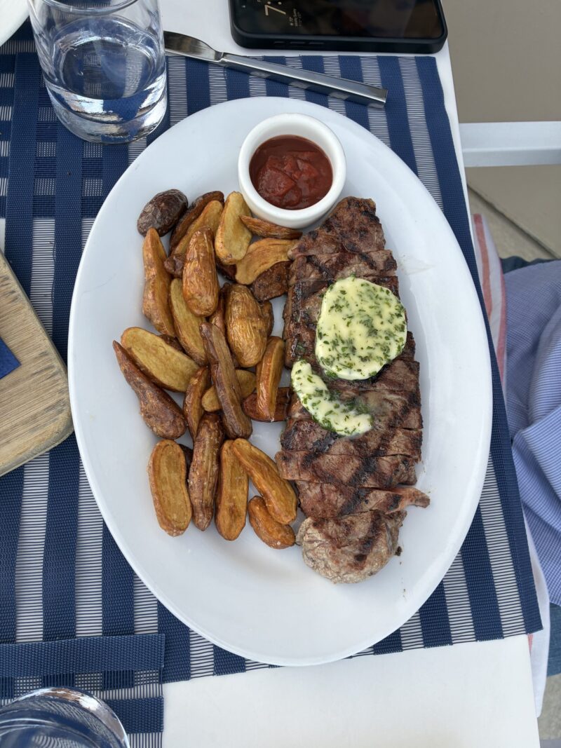 Grilled NY strip steak topped with herb butter, served with roasted fingerling potatoes and ketchup on a white platter at Carbon Beach Club Restaurant at the Malibu Beach Inn
