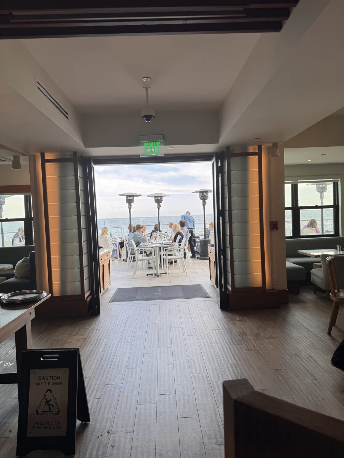 Interior view of Carbon Beach Club Restaurant at the Malibu Beach Inn, looking through open doors to an oceanfront patio where guests dine at white tables beside tall heaters, with the Pacific Ocean beyond