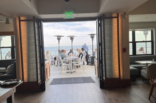 Interior view of Carbon Beach Club Restaurant at the Malibu Beach Inn, looking through open doors to an oceanfront patio where guests dine at white tables beside tall heaters, with the Pacific Ocean beyond
