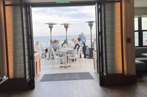 Interior view of Carbon Beach Club Restaurant at the Malibu Beach Inn, looking through open doors to an oceanfront patio where guests dine at white tables beside tall heaters, with the Pacific Ocean beyond