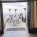 Interior view of Carbon Beach Club Restaurant at the Malibu Beach Inn, looking through open doors to an oceanfront patio where guests dine at white tables beside tall heaters, with the Pacific Ocean beyond
