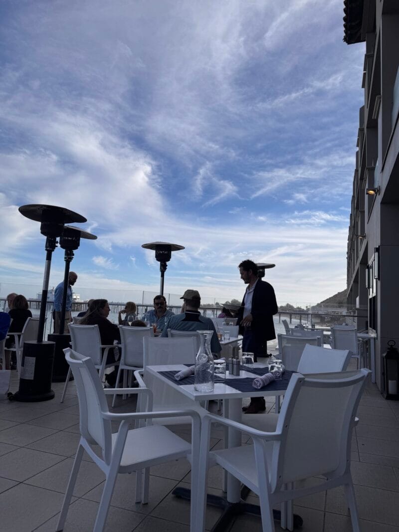 The oceanfront patio at Carbon Beach Club Restaurant at the Malibu Beach Inn, with white tables and chairs, tall patio heaters, and diners overlooking the Pacific Ocean and coastline under a blue sky