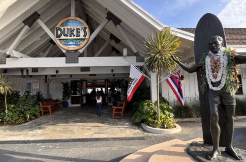 The exterior entrance of Duke's Malibu restaurant, with its peaked A-frame roof and round Duke's sign, a bronze statue of surfer Duke Kahanamoku draped in flower leis, and Hawaiian and California flags under a blue sky