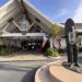 The exterior entrance of Duke's Malibu restaurant, with its peaked A-frame roof and round Duke's sign, a bronze statue of surfer Duke Kahanamoku draped in flower leis, and Hawaiian and California flags under a blue sky