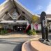 The exterior entrance of Duke's Malibu restaurant, with its peaked A-frame roof and round Duke's sign, a bronze statue of surfer Duke Kahanamoku draped in flower leis, and Hawaiian and California flags under a blue sky
