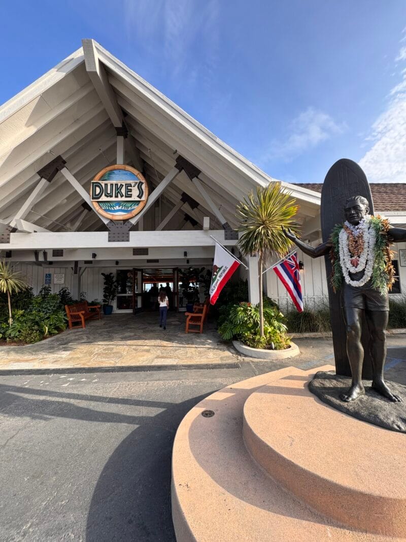 The exterior entrance of Duke's Malibu restaurant, with its peaked A-frame roof and round Duke's sign, a bronze statue of surfer Duke Kahanamoku draped in flower leis, and Hawaiian and California flags under a blue sky