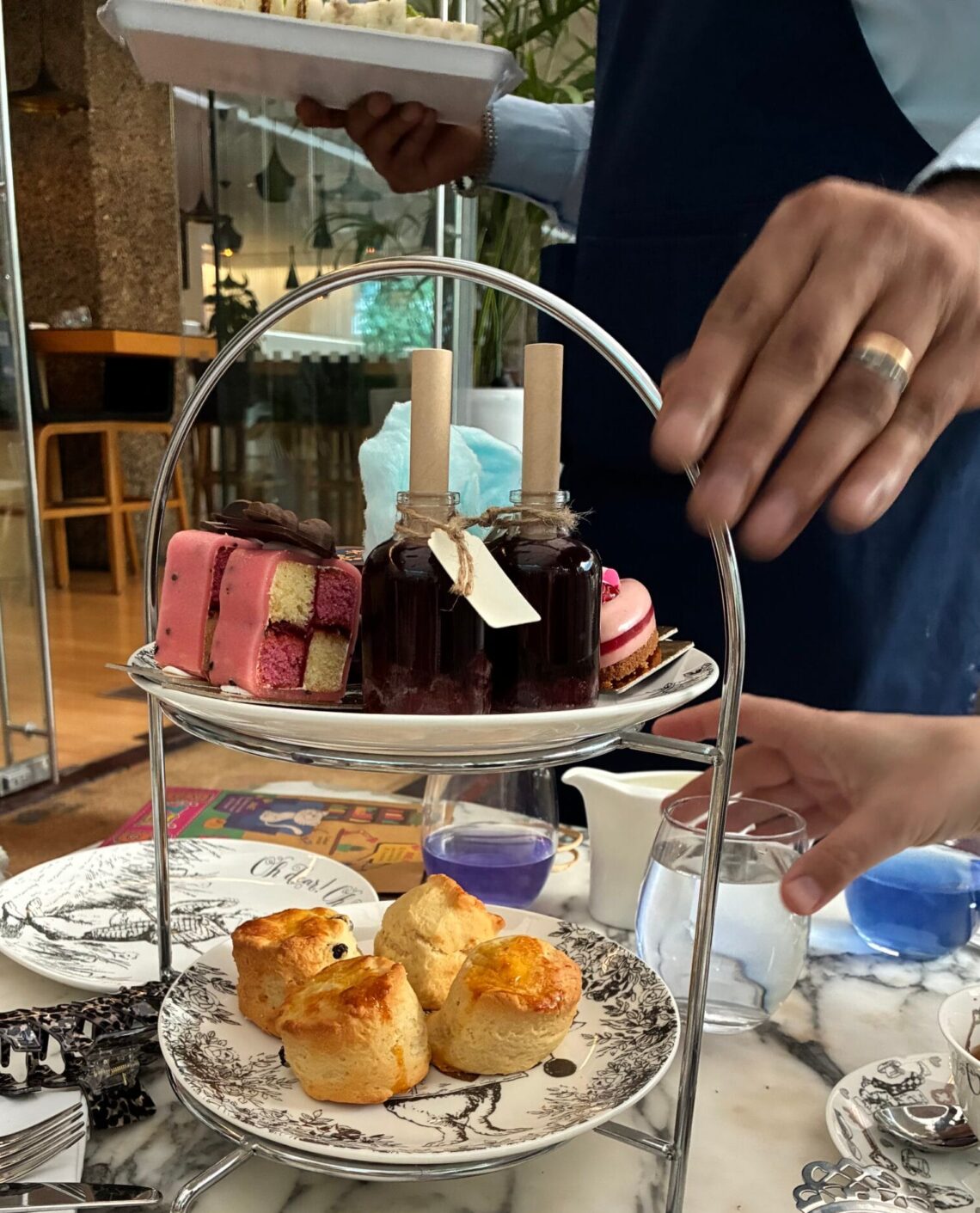 A tiered stand of Mad Hatters Afternoon Tea treats, featuring freshly baked scones on illustrated china, colorful desserts, and two bottled “Drink Me” potions, with a server presenting tea sandwiches in the background.