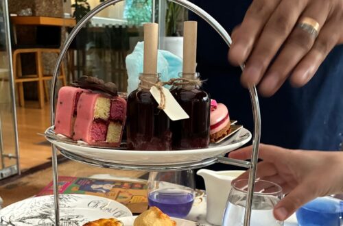A tiered stand of Mad Hatters Afternoon Tea treats, featuring freshly baked scones on illustrated china, colorful desserts, and two bottled “Drink Me” potions, with a server presenting tea sandwiches in the background.