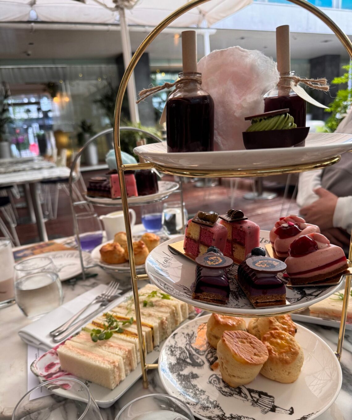 A tiered stand of Mad Hatters Afternoon Tea treats, including scones, colorful pastries, chocolate desserts, “Drink Me” potion bottles, and pink cotton candy, with tea sandwiches arranged on a plate beside it.