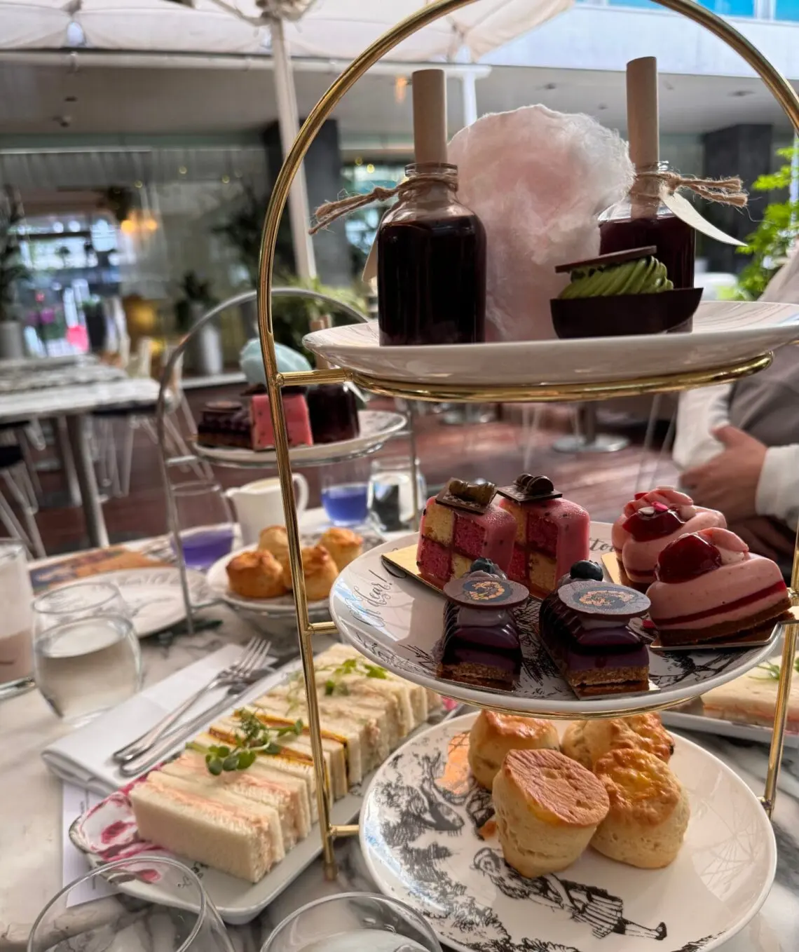 A tiered stand of Mad Hatters Afternoon Tea treats, including scones, colorful pastries, chocolate desserts, “Drink Me” potion bottles, and pink cotton candy, with tea sandwiches arranged on a plate beside it.