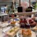 A tiered stand of Mad Hatters Afternoon Tea treats, including scones, colorful pastries, chocolate desserts, “Drink Me” potion bottles, and pink cotton candy, with tea sandwiches arranged on a plate beside it.