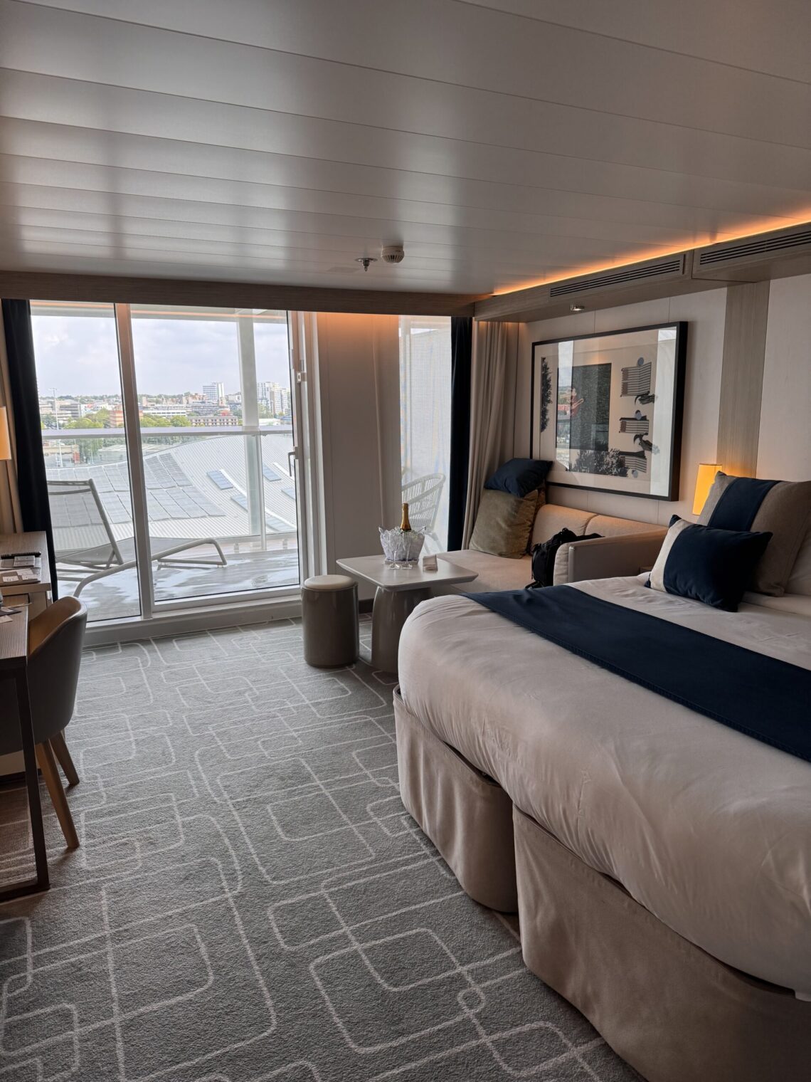 Our Sky Suite aboard Celebrity Apex featuring a king-size bed with white bedding and navy accents, a sitting area, desk, and floor-to-ceiling glass doors opening to a private balcony with outdoor loungers and a city view.