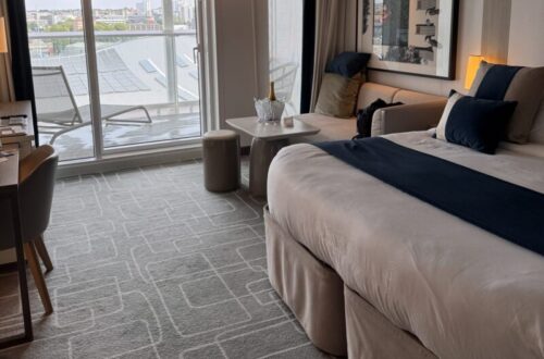 Our Sky Suite aboard Celebrity Apex featuring a king-size bed with white bedding and navy accents, a sitting area, desk, and floor-to-ceiling glass doors opening to a private balcony with outdoor loungers and a city view.