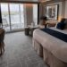 Our Sky Suite aboard Celebrity Apex featuring a king-size bed with white bedding and navy accents, a sitting area, desk, and floor-to-ceiling glass doors opening to a private balcony with outdoor loungers and a city view.