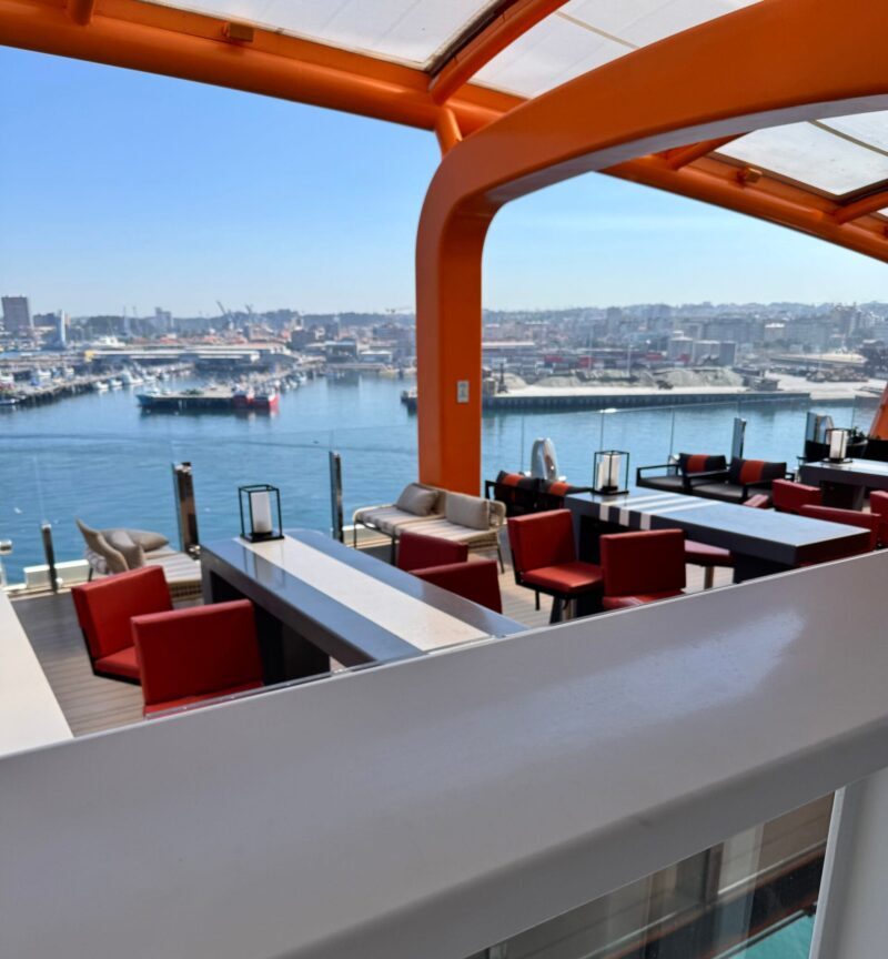The orange Magic Carpet platform on Celebrity Apex with red chairs and a harbor view