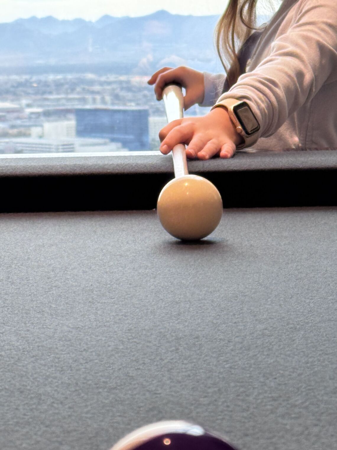 A child's hand lines up a shot on the pool table in our suite at the Venetian, with the Las Vegas skyline and mountains visible through the window