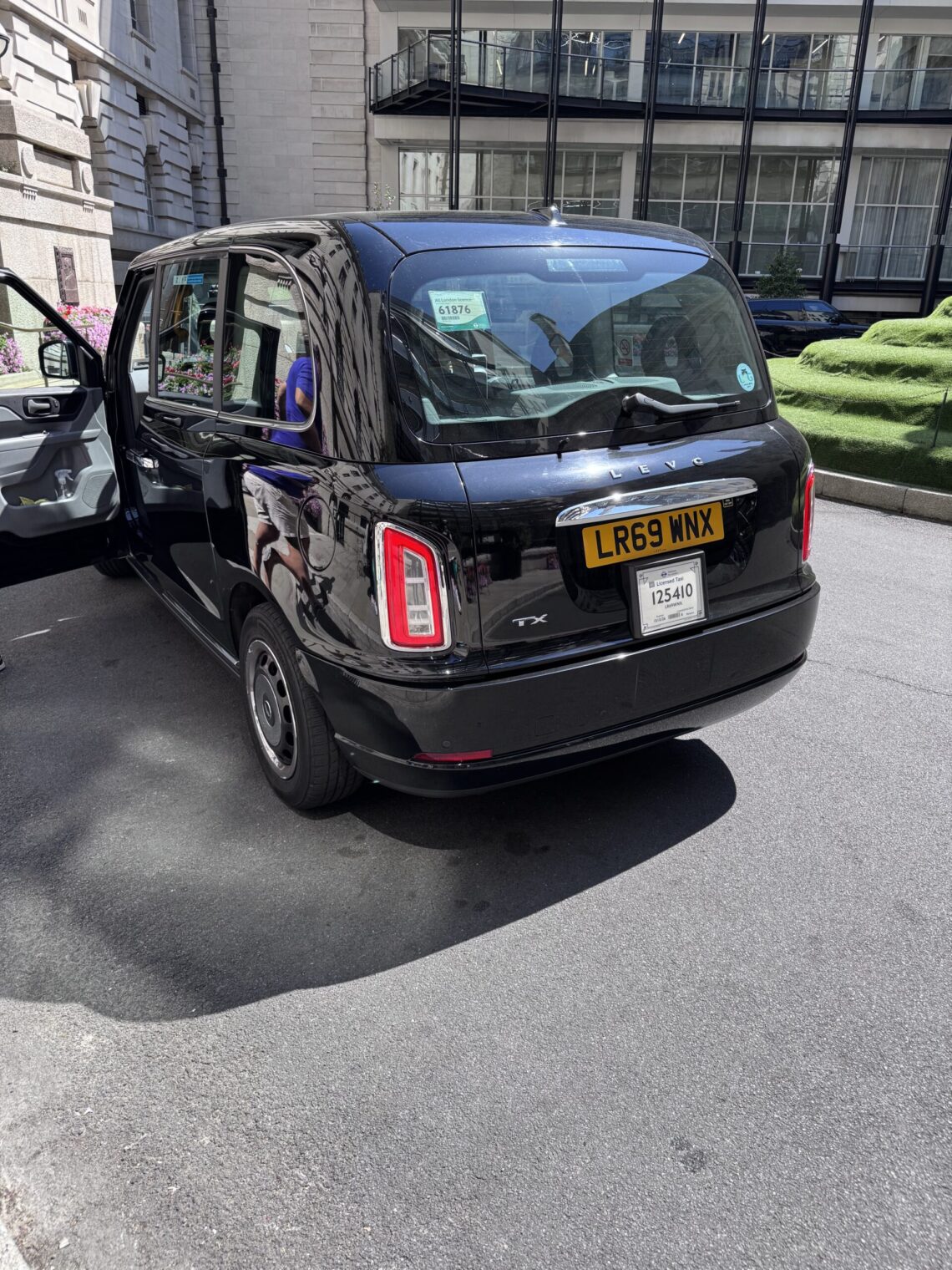A black London taxi parked outside our hotel in London, showing its distinctive boxy shape, glossy finish, and yellow rear license plate.