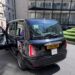 A black London taxi parked outside our hotel in London, showing its distinctive boxy shape, glossy finish, and yellow rear license plate.