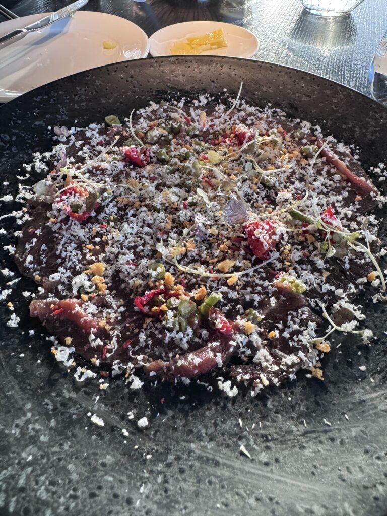 Dry-aged Irish beef carpaccio from the Aqua Shard Sunset Menu, served on a black plate with grated white garnish, herbs, microgreens, red vegetables, and crunchy crumbs.