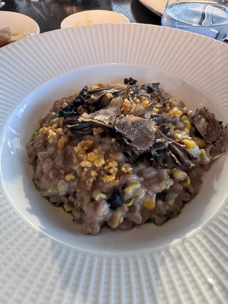 A bowl of creamy sweetcorn and mushroom spelt risotto topped with crispy grains, mushrooms, herbs, and thin shavings of truffle, served on a white ridged plate.