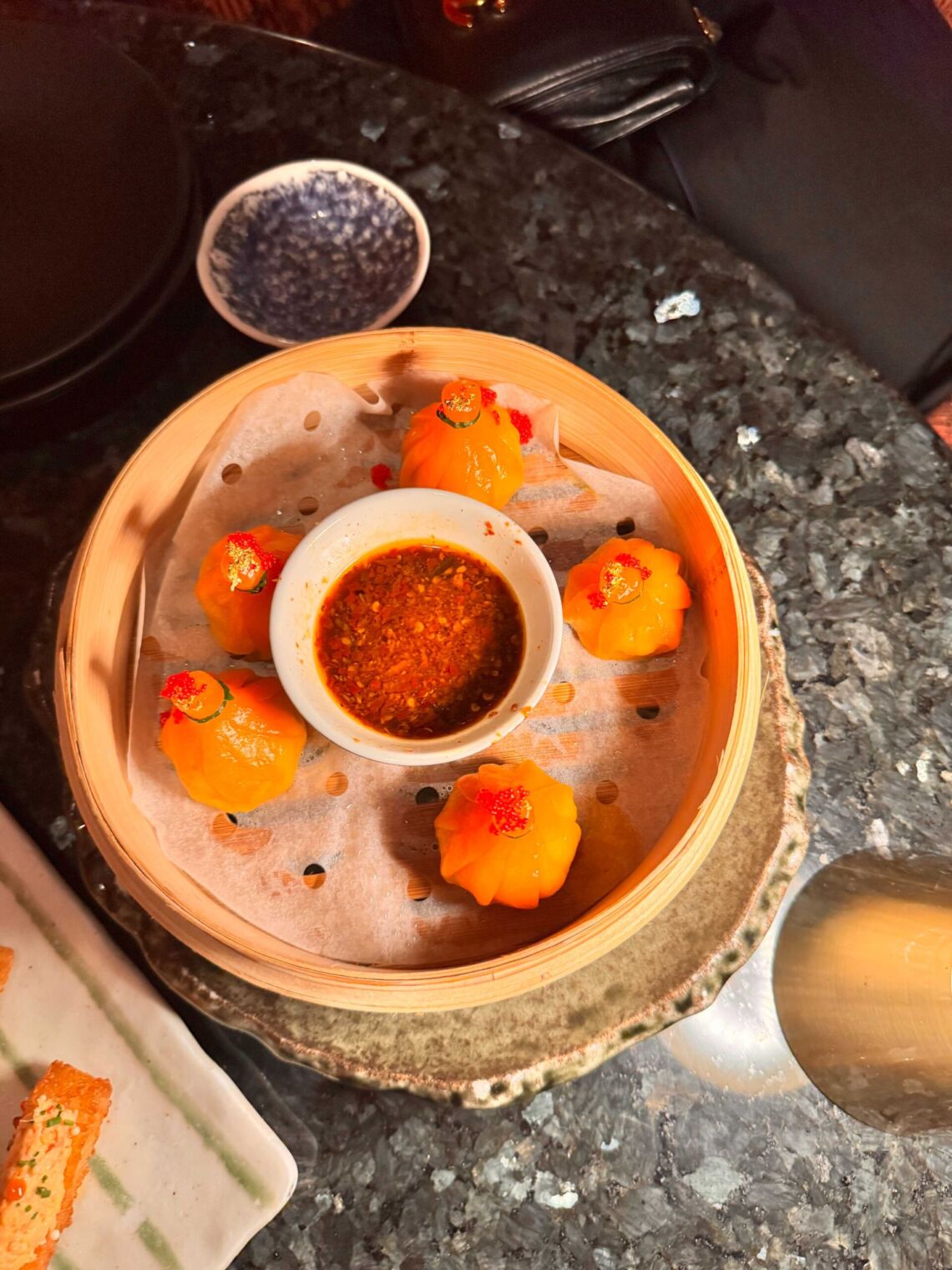 Bamboo steamer basket of orange dumplings topped with red tobiko around a bowl of chili oil dipping sauce