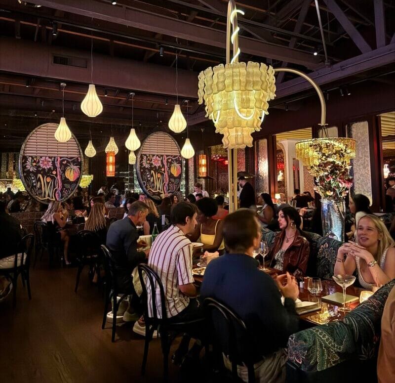 The busy main dining room at Bad Roman Beverly Hills with lantern lighting, a large chandelier, and diners at full tables.