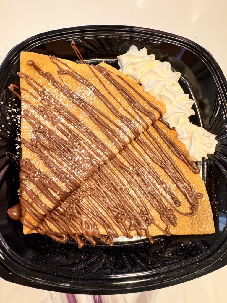 A folded Nutella crepe drizzled with chocolate sauce and topped with whipped cream, on a black plate