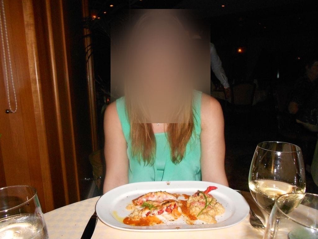 A plated lobster tail with risotto and a roasted red pepper at a cruise ship specialty restaurant, served with a glass of white wine.