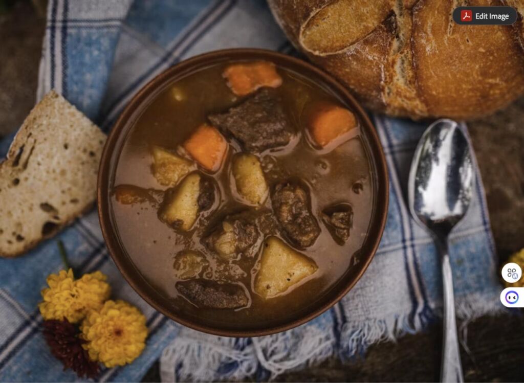 A bowl of hearty beef stew with potatoes and carrots, served with crusty bread, a spoon, and marigold flowers on a plaid cloth.