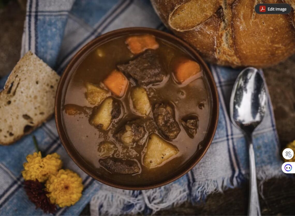 A bowl of hearty beef stew with potatoes and carrots, served with crusty bread, a spoon, and marigold flowers on a plaid cloth.