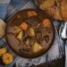 A bowl of hearty beef stew with potatoes and carrots, served with crusty bread, a spoon, and marigold flowers on a plaid cloth.