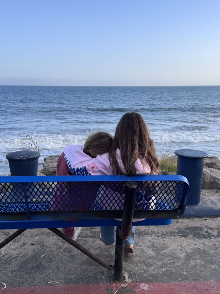 Two girls sitting on a bench with their arms around each other, looking out at the ocean at Will Rogers State Beach near Gladstones Malibu.