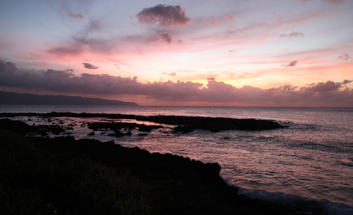 Pink and orange sunset over the North Shore coastline near Turtle Bay, Oahu, with dark lava rock in the foreground — the view near Alaia restaurant at dinner time.