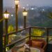 A rooftop lounge with wicker chairs and string lights overlooking a hazy city skyline at dusk, evoking upscale Los Angeles rooftop dining