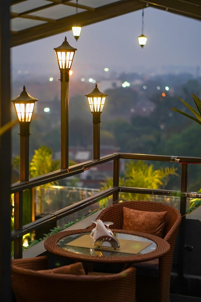 A rooftop lounge with wicker chairs and string lights overlooking a hazy city skyline at dusk, evoking upscale Los Angeles rooftop dining