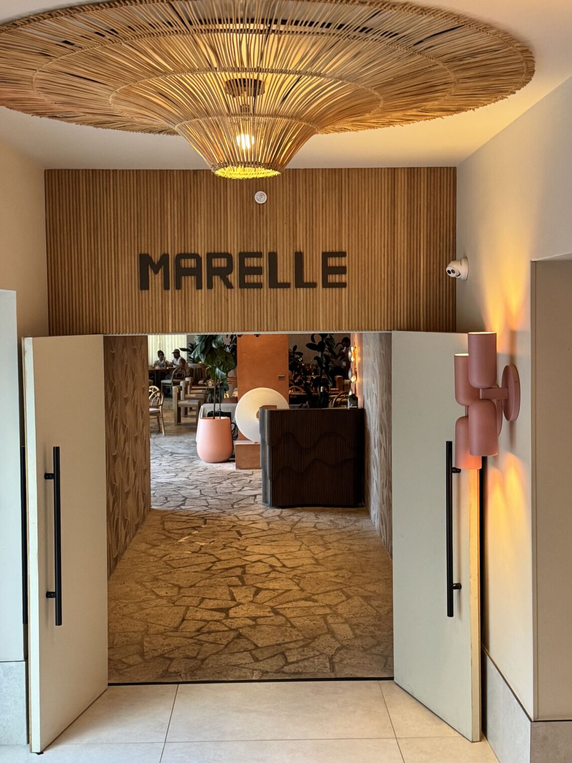 The entrance to Marelle in Santa Monica, with the restaurant name on a wood-paneled wall, a woven pendant light overhead, and open doors leading to a stone-floored dining room