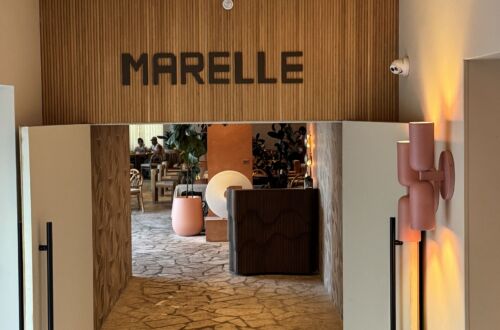The entrance to Marelle in Santa Monica, with the restaurant name on a wood-paneled wall, a woven pendant light overhead, and open doors leading to a stone-floored dining room