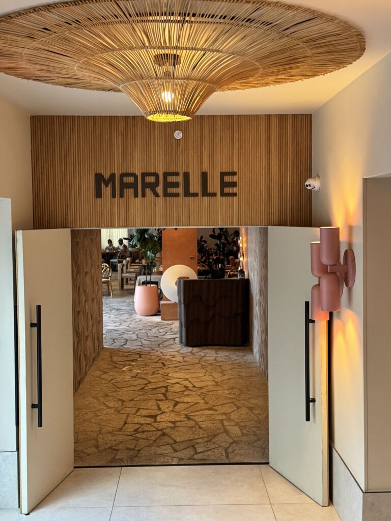The entrance to Marelle in Santa Monica, with the restaurant name on a wood-paneled wall, a woven pendant light overhead, and open doors leading to a stone-floored dining room