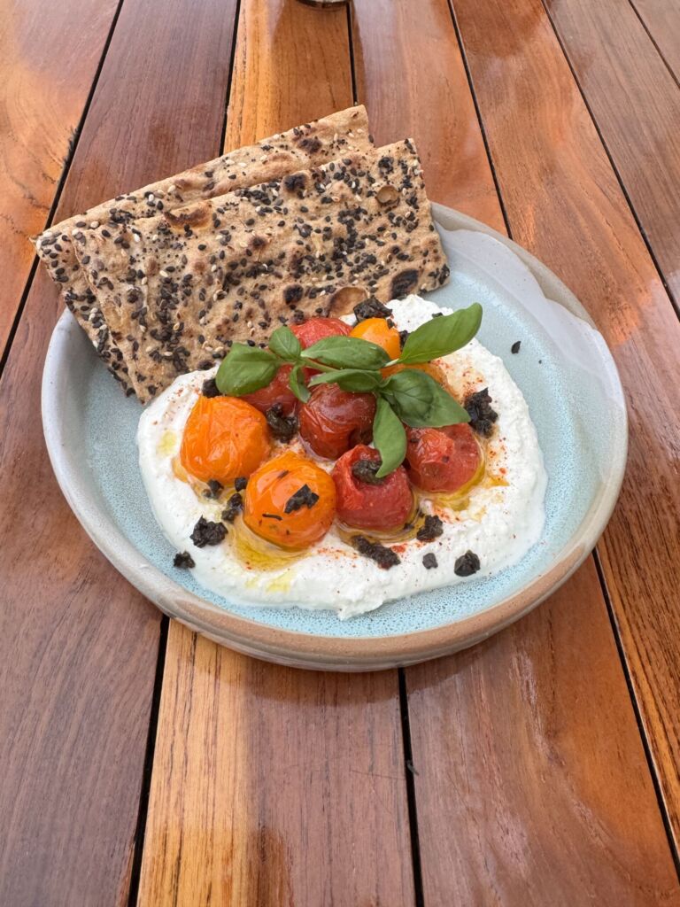 Whipped feta at Marelle topped with roasted cherry tomatoes, fresh basil, and black olive crumble, served with seeded crispbread on a blue plate
