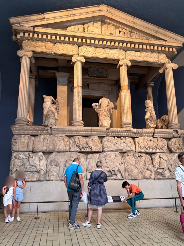 A detailed view of the Nereid Monument, an ancient Greek marble temple structure on display at the British Museum, featuring fluted columns, intricate friezes, and headless statues of female figures positioned between the pillars.