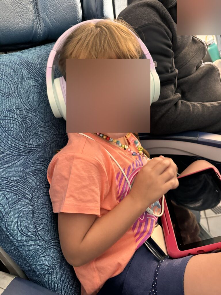 Our daughter in a Hawaiian Airlines seat with headphones and a tablet, face blurred