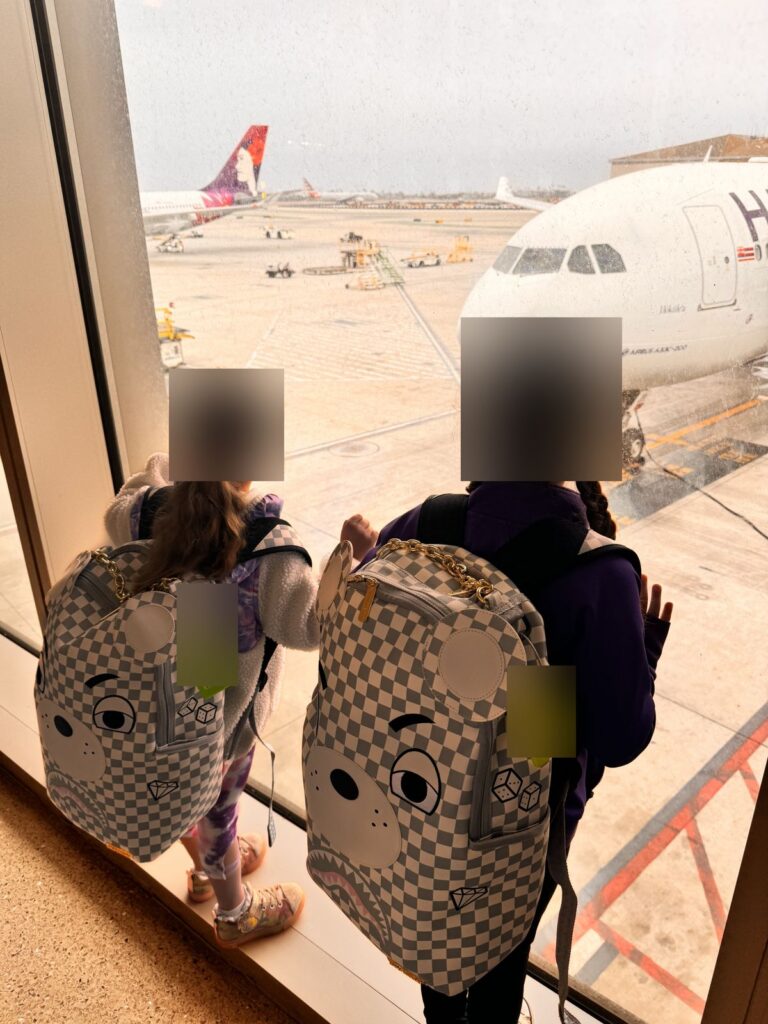 Our daughters at the LAX gate window watching the Hawaiian Airlines plane, seen from behind and blurred