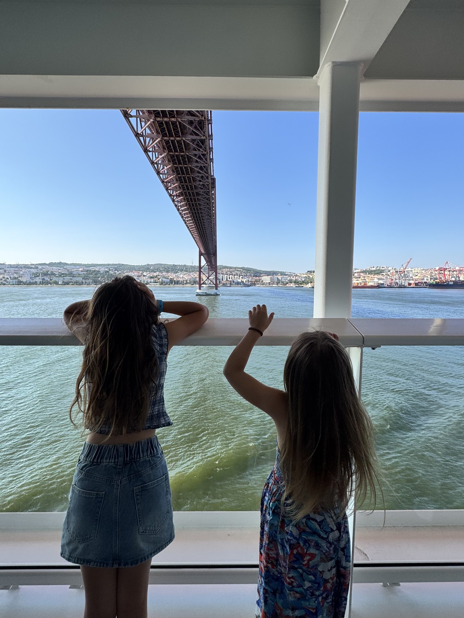 Two girls on a cruise ship deck looking up at the 25 de Abril Bridge in Lisbon